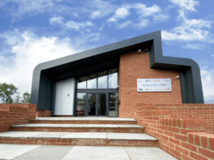 Contemporary new build office building, designed by a Norwich-based architect. This striking design features a combination of traditional red brickwork and bold modern detailing, including a sculptural steel-framed roofline. The entrance is highlighted by large glazed panels, allowing natural light to flood the interior, and a welcoming stepped pathway framed by brick landscaping. The building demonstrates a balance of functionality and cutting-edge architectural design.
