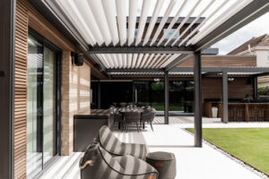 Contemporary new build home in Norfolk, designed by a Norwich-based architect. This modern residence features an expansive outdoor living area with a sleek brise soleil shading system, offering both style and functional solar control. The design integrates timber cladding, brickwork, and floor-to-ceiling glazing, seamlessly connecting the indoor and outdoor spaces. An outdoor kitchen and dining area enhance the home's focus on modern, open-plan living, while the carefully selected materials emphasize a sophisticated yet natural palette.
