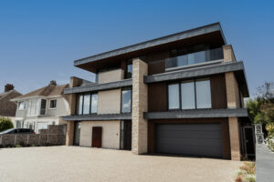 Contemporary new build home in Norfolk, designed by a Norwich-based architect. This striking three-story residence showcases a bold modern aesthetic with flat roofs, zinc and timber cladding, and large glazed openings. The design incorporates gabion walls, natural stone accents, and recessed balconies for a refined architectural statement. The use of sustainable materials and a minimalist palette emphasizes clean lines and a strong connection to its coastal setting.