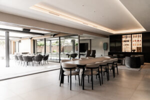 Contemporary new build home in Norfolk, designed by a Norwich-based architect. The open-plan living area seamlessly connects to the outdoor patio through large sliding glass doors, enhancing the sense of space and natural light. Modern finishes include a bespoke bar with backlit shelving, minimalist furniture, and integrated lighting. The design emphasizes a fluid connection between indoor and outdoor spaces, perfect for modern family living and entertaining.